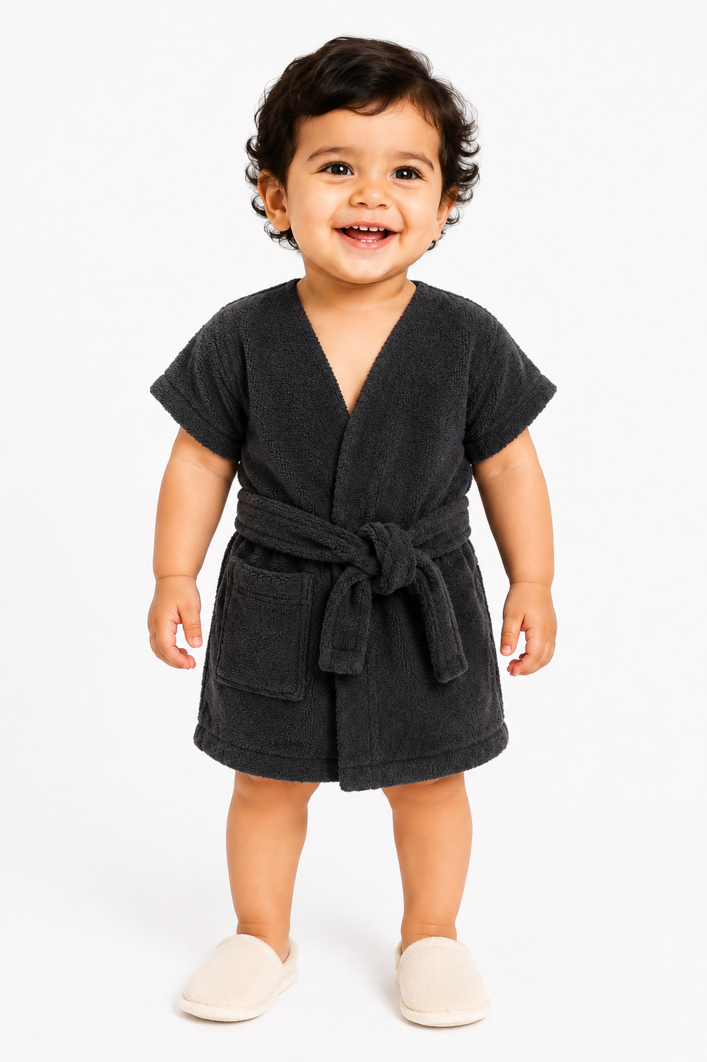 A toddler standing against a white studio background, shown in two views wearing a dark charcoal gray short-sleeve bathrobe with a tie belt and front patch pockets. The child is also wearing off-white slippers.