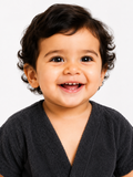 A close-up portrait of a smiling toddler with dark curly hair wearing a dark gray textured V-neck top, set against a plain light gray background.