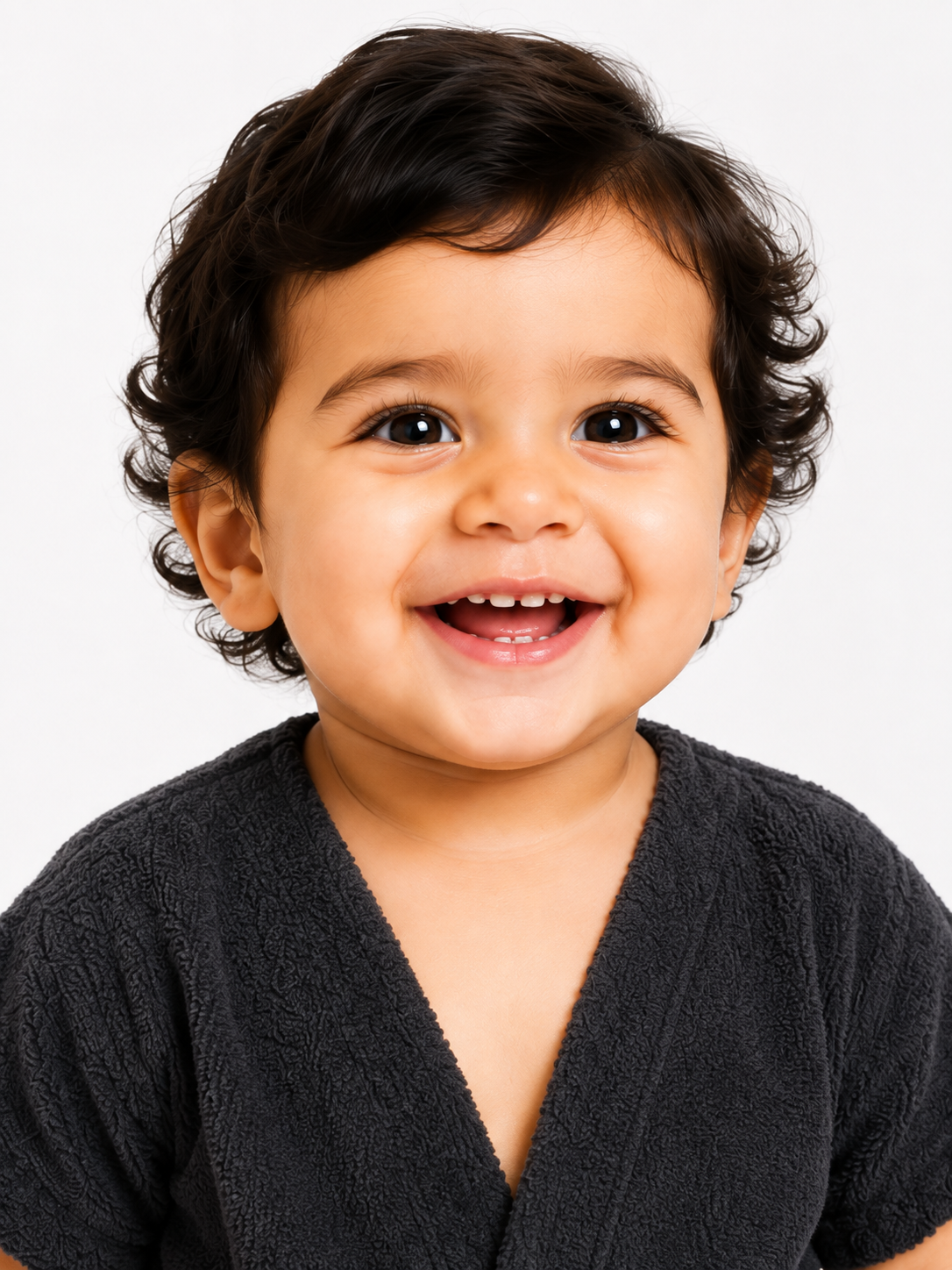 A close-up portrait of a smiling toddler with dark curly hair wearing a dark gray textured V-neck top, set against a plain light gray background.
