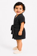 A smiling toddler standing in profile wearing a dark charcoal gray terry cloth bathrobe with a tie belt and light beige slip-on slippers, photographed against a seamless white studio background.