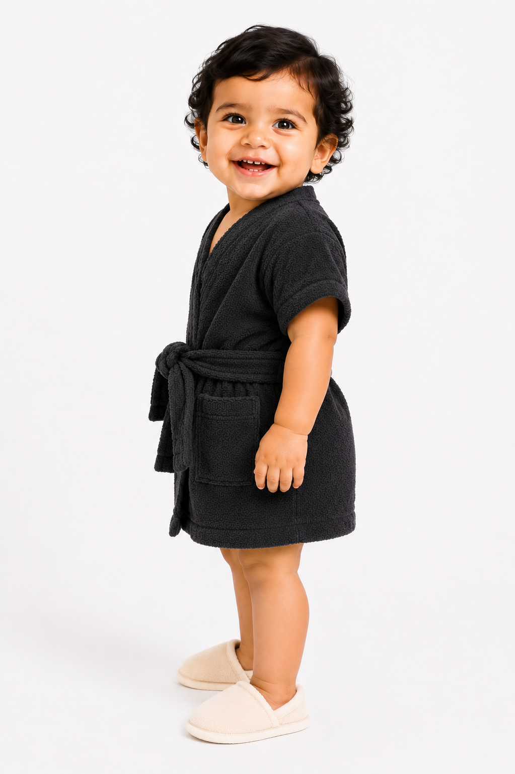 A smiling toddler standing in profile wearing a dark charcoal gray terry cloth bathrobe with a tie belt and light beige slip-on slippers, photographed against a seamless white studio background.