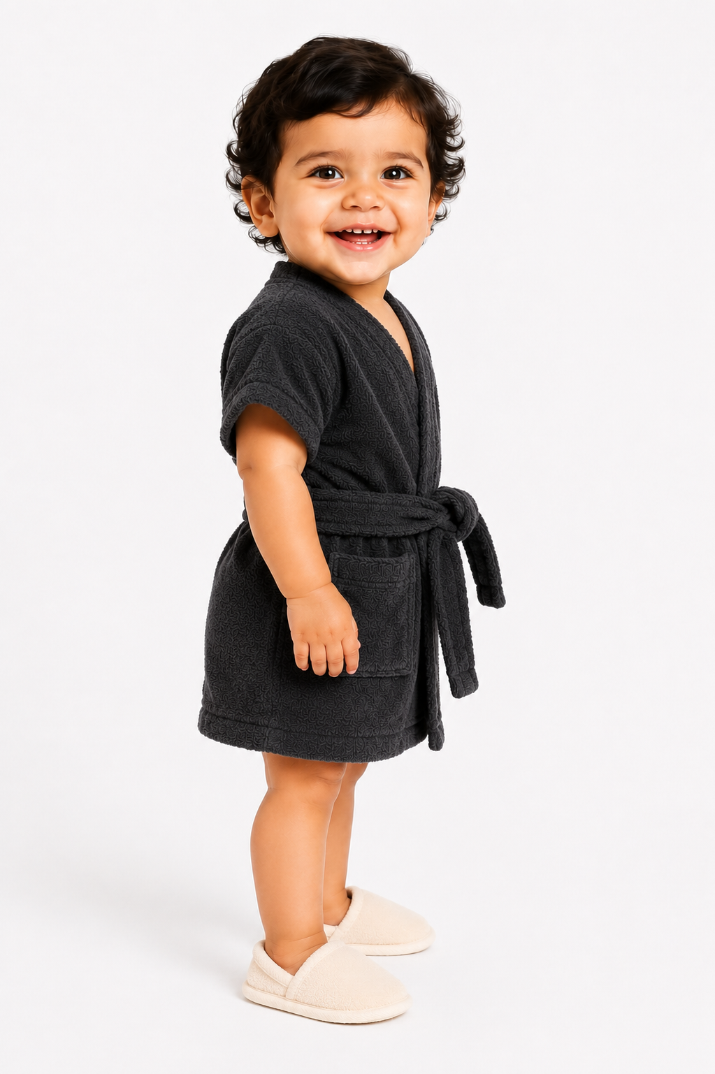 A smiling toddler standing against a light gray studio background, wearing a dark charcoal gray terry cloth bathrobe with a tie belt and light beige slip-on slippers.
