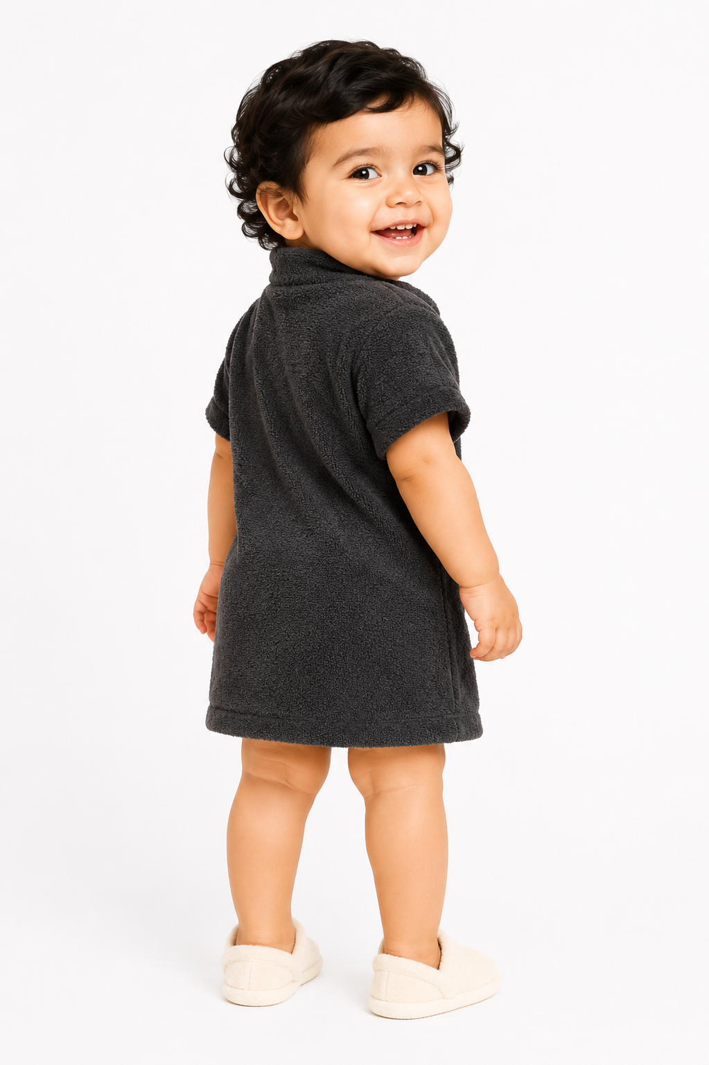 A toddler with curly dark hair is shown from the back wearing a dark charcoal gray short-sleeve terry cloth dress or romper, paired with cream-colored slip-on shoes, against a plain white studio background.