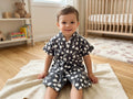 A young child sitting on a cream towel in a bright nursery, wearing a dark gray short-sleeve robe with a white cloud pattern and the word 'LOVE' printed on it.