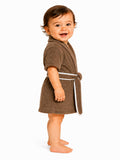 A smiling toddler standing barefoot in profile, wearing a brown short-sleeve bathrobe with white piping and a matching tie belt, photographed against a clean white background.