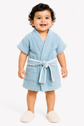 A smiling toddler wearing a light blue terry cloth bathrobe with a white tie belt, standing against a plain white studio background.