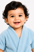 A close-up of a smiling toddler with dark curly hair wearing a light blue textured bathrobe against a plain white background.