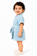 A smiling toddler standing in profile wearing a light blue terry cloth bathrobe with a tie belt and white slip-on shoes, photographed against a clean white studio background.