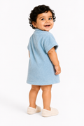 A toddler with curly dark hair is shown from the back wearing a light blue short-sleeve terry cloth dress or romper, paired with cream-colored slip-on shoes, against a plain white studio background.
