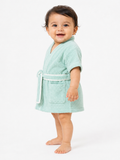 A smiling toddler standing barefoot against a plain white studio background, wearing a short mint green terry cloth bathrobe with a white belt and front patch pocket.