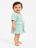 A smiling toddler standing barefoot against a plain white studio background, wearing a short-sleeve mint green terry cloth bathrobe with a white belt and front patch pocket.
