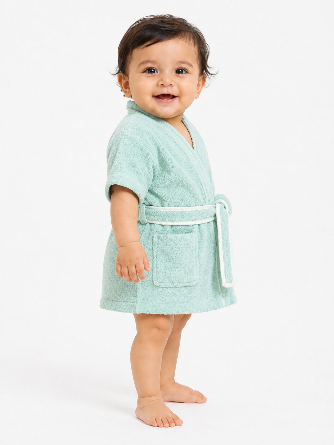 A smiling toddler standing barefoot against a plain white studio background, wearing a short-sleeve mint green terry cloth bathrobe with a white belt and front patch pocket.