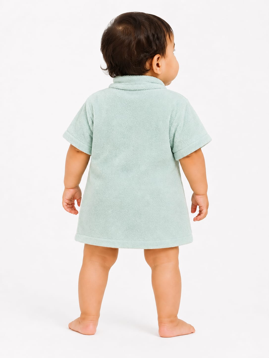 A baby standing from the back wearing a light mint green short-sleeve t-shirt dress, photographed against a clean white studio background.