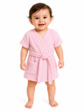 A smiling baby standing barefoot on a white background, wearing a soft pink short-sleeve terry cloth bathrobe with a tie belt and front patch pocket.
