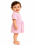 A smiling baby standing in profile wearing a soft pink terry cloth bathrobe with a tie belt and front pocket, photographed against a clean white studio background.