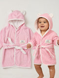 A baby wearing a pink hooded bathrobe with bear ears and a white lining, standing next to a matching pink bathrobe hanging flat against a light gray background.
