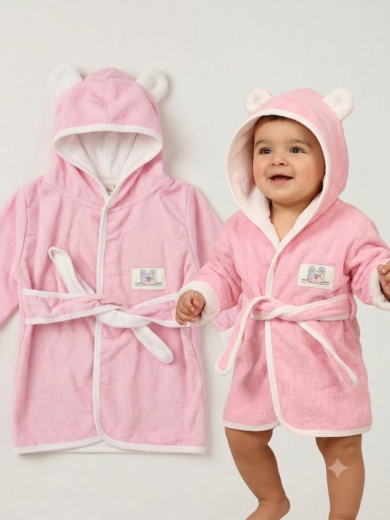 A baby wearing a pink hooded bathrobe with bear ears and a white lining, standing next to a matching pink bathrobe hanging flat against a light gray background.