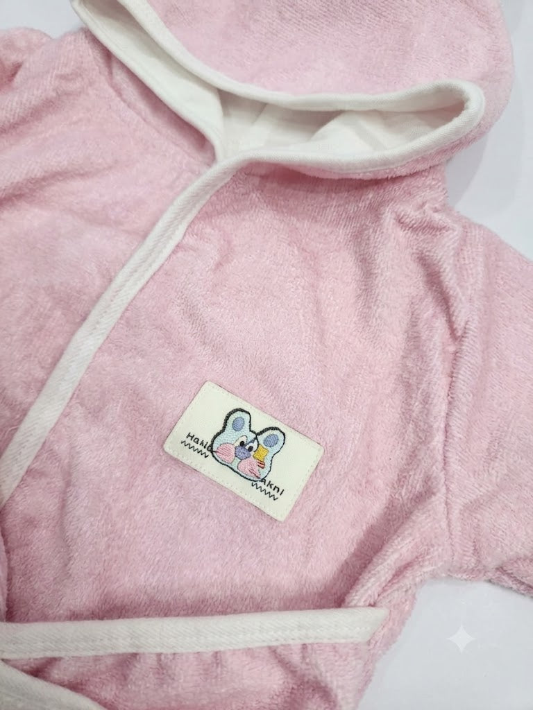 A close-up of a pink plush hooded towel robe with cream-colored trim and a small embroidered patch featuring a cartoon mouse and the text 'HABIBI GABONNI'.