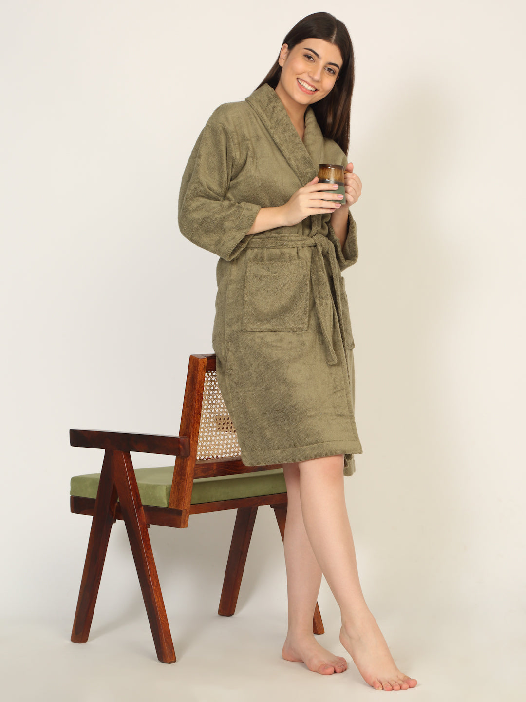 Woman wearing a green robe holding a glass, standing next to a wooden chair with a white background