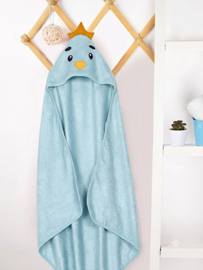 A light blue hooded towel with a cartoon bird face and small gold crown, hanging on a wooden wall rack in a bright indoor setting.