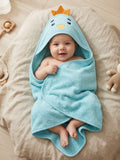 A baby wrapped in a light blue hooded towel with a cartoon bird face and small yellow crown, lying on a beige cushion with toys nearby.