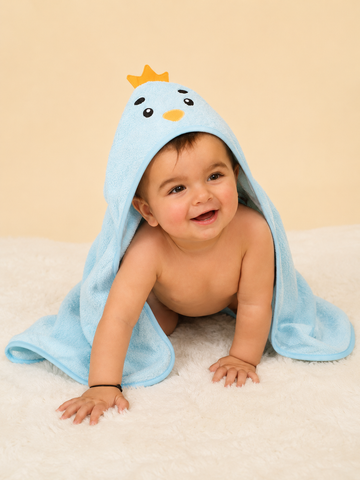 A baby crawling on a white textured blanket, wrapped in a light blue hooded towel with a cartoon bird face and small yellow crown detail.