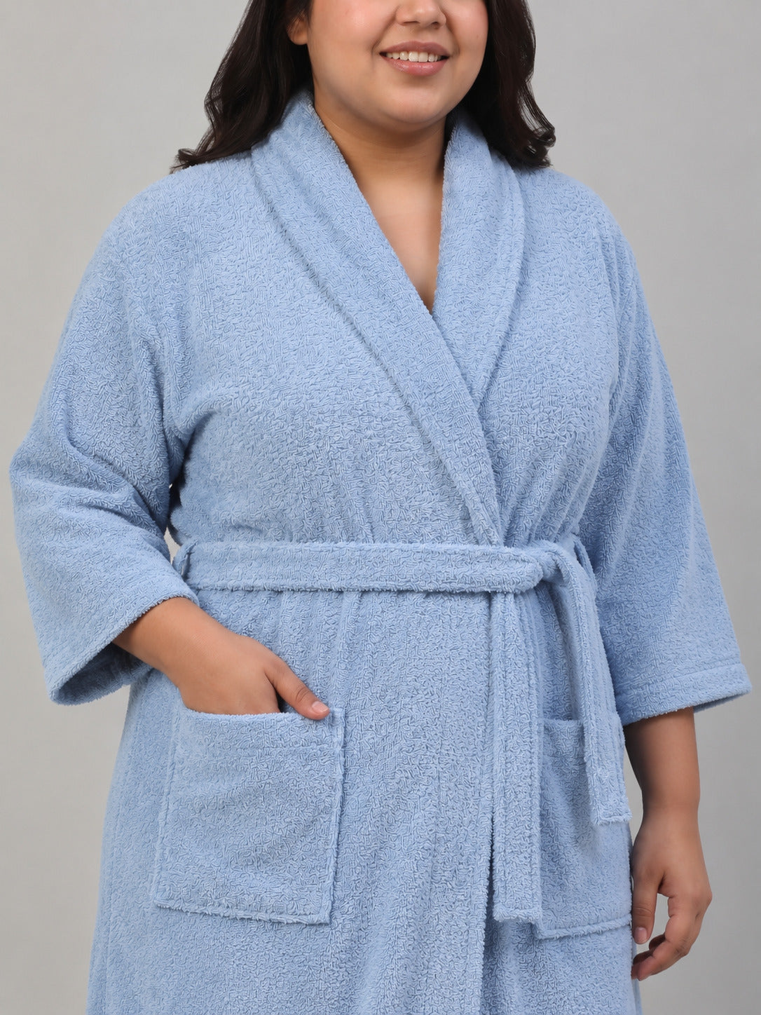 A person wearing a light blue terry cloth bathrobe with a shawl collar, self-tie belt, and front patch pockets, photographed against a plain light gray studio background.
