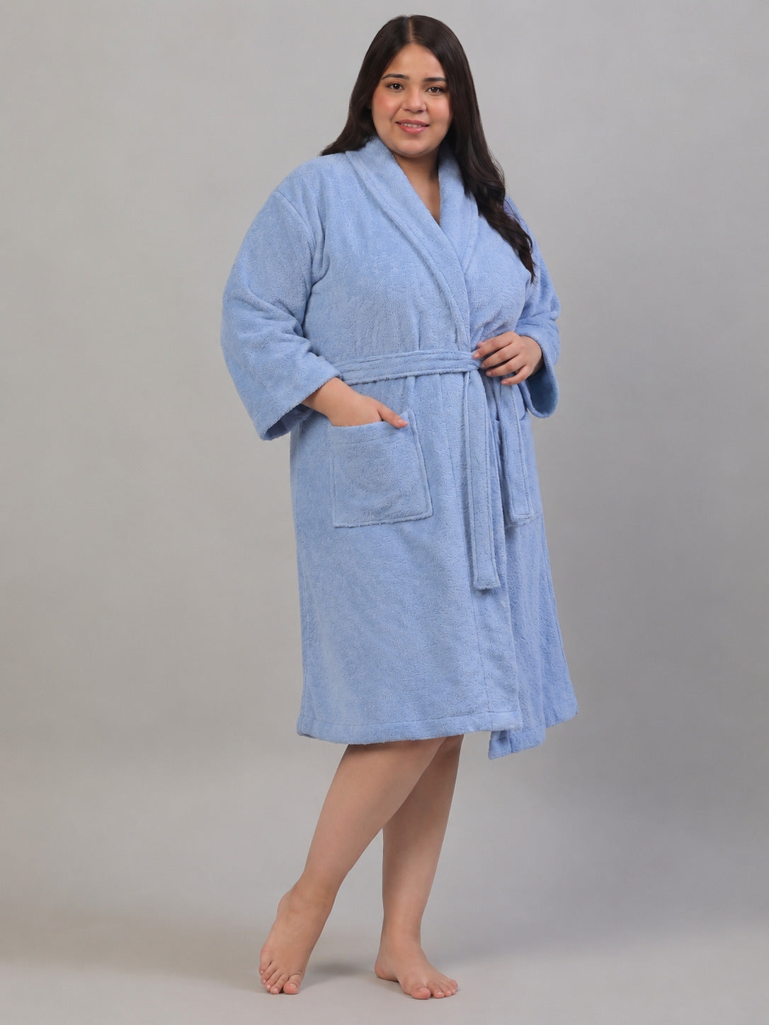 A woman modeling a light blue terry cloth bathrobe with a shawl collar, front patch pocket, and self-tie belt, standing against a neutral gray studio background.