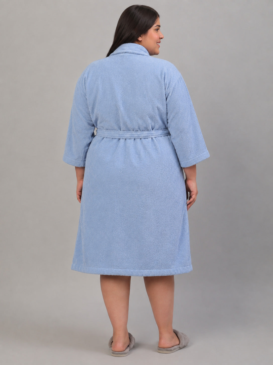 A woman wearing a light blue terry cloth bathrobe, shown from the back against a solid gray studio background.