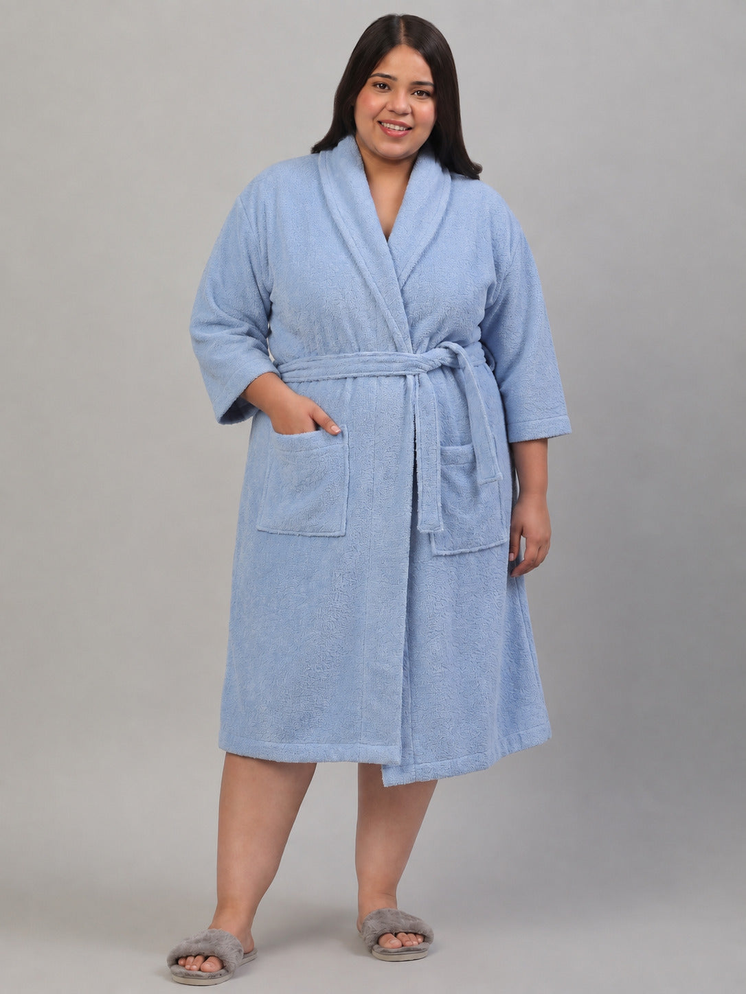 A woman wearing a light blue textured bathrobe stands next to a dark wooden chair with a beige cushion, holding an open book. She is wearing gray slippers against a solid gray studio background.