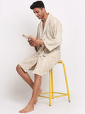 A man wearing a cream-colored textured bathrobe sits on a yellow stool, reading a book against a plain white background.