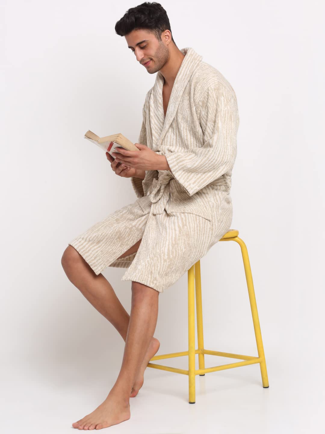 A man wearing a cream-colored textured bathrobe sits on a yellow stool, reading a book against a plain white background.