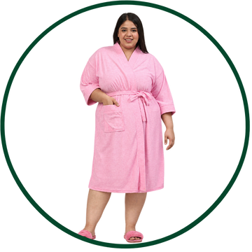 A woman modeling a pink terry cloth bathrobe with a tie waist and front patch pocket, paired with pink slip-on slippers, set against a white background with a dark green circular border.
