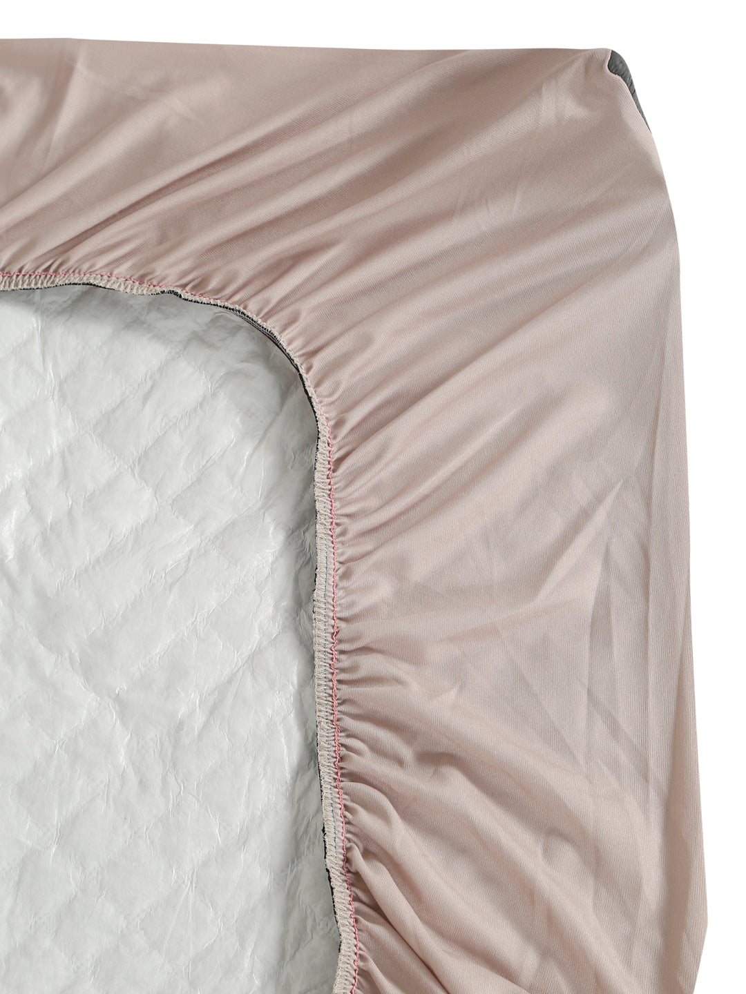 A close-up of a fitted sheet with gathered elastic edges, shown partially pulled over a mattress. The sheet is a dusty blush pink color, and the mattress underneath is light gray.