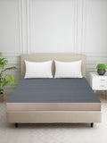 A dark gray mattress topper or fitted sheet on a bed frame with two white pillows, set in a modern bedroom with a white paneled wall, a side table, and a plant.