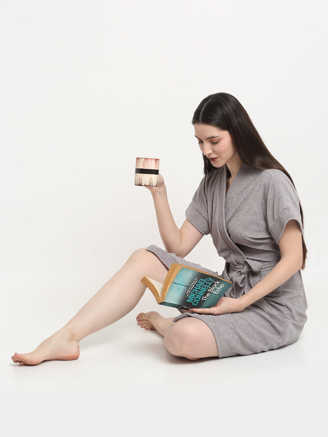 A woman sitting and wearing a grey micro terry bathrobe and holding a book and a cup