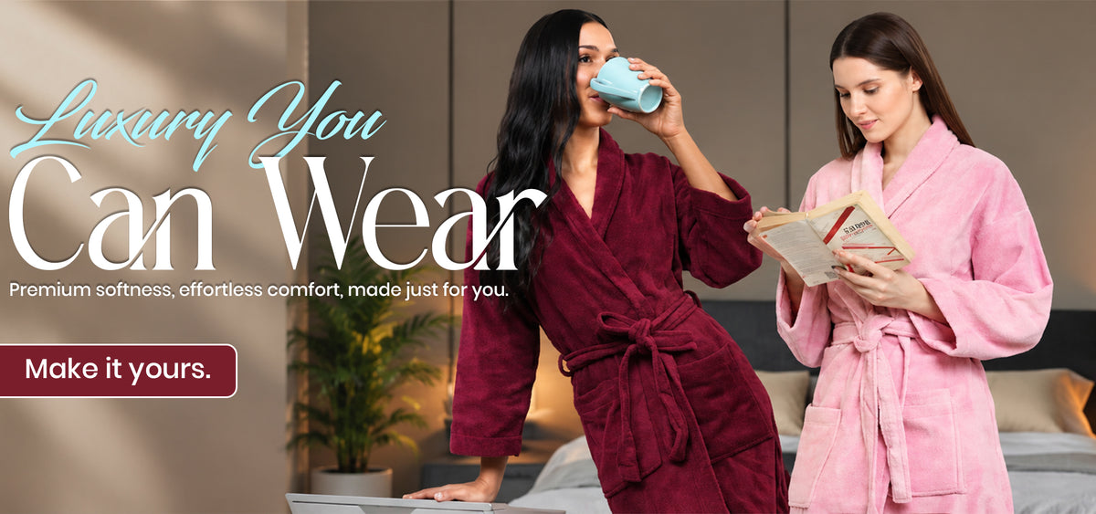 Two women in plush bathrobes in a home-like setting. One wears a deep burgundy robe and drinks from a light blue mug, while the other wears a pink robe and reads a book.1621243260e1af0c20-0