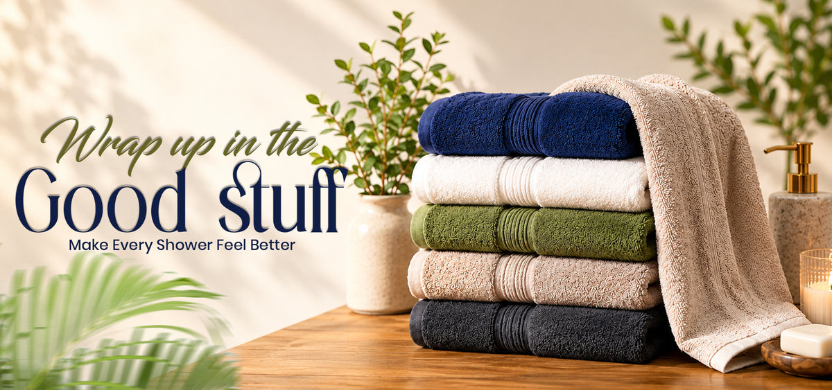A stack of five folded bath towels in navy, cream, olive green, beige, and charcoal gray, with a beige towel draped over the side. The scene is styled on a wooden surface with a vase of greenery and a soap dispenser in the background, with the text 'Wrap up in the Good Stuff' and 'Make Every Shower Feel Better' overlaid at the top.slider_item_ecNkVL