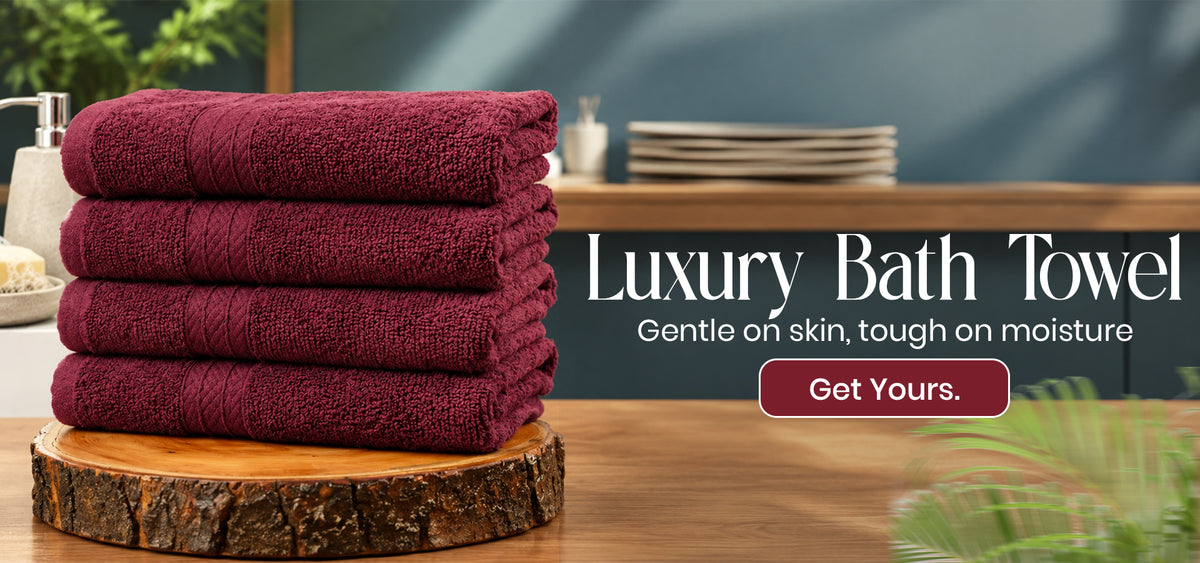 A stack of four folded bath towels in a deep burgundy color, displayed on a rustic wooden log slice. The background is a softly blurred bathroom setting with green foliage and a dark teal wall, featuring promotional text 'Luxury Bath Towel' and a 'Get Yours' button.slider_item_WkPKyE