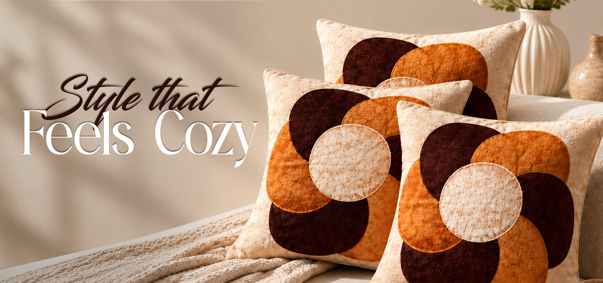 Three decorative throw pillows with a retro circular patchwork pattern in beige, orange, and dark brown, styled on a beige sofa with a textured blanket. The text 'Style that Feels Cozy' is displayed on the left side.slider_item_V6zBdB