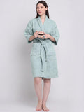 A woman wearing a light sage green bathrobe, holding a small cup, standing against a plain light gray studio background.