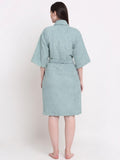 A woman viewed from the back wearing a short-sleeved, belted bathrobe in a muted sage green color, standing against a plain white studio background.