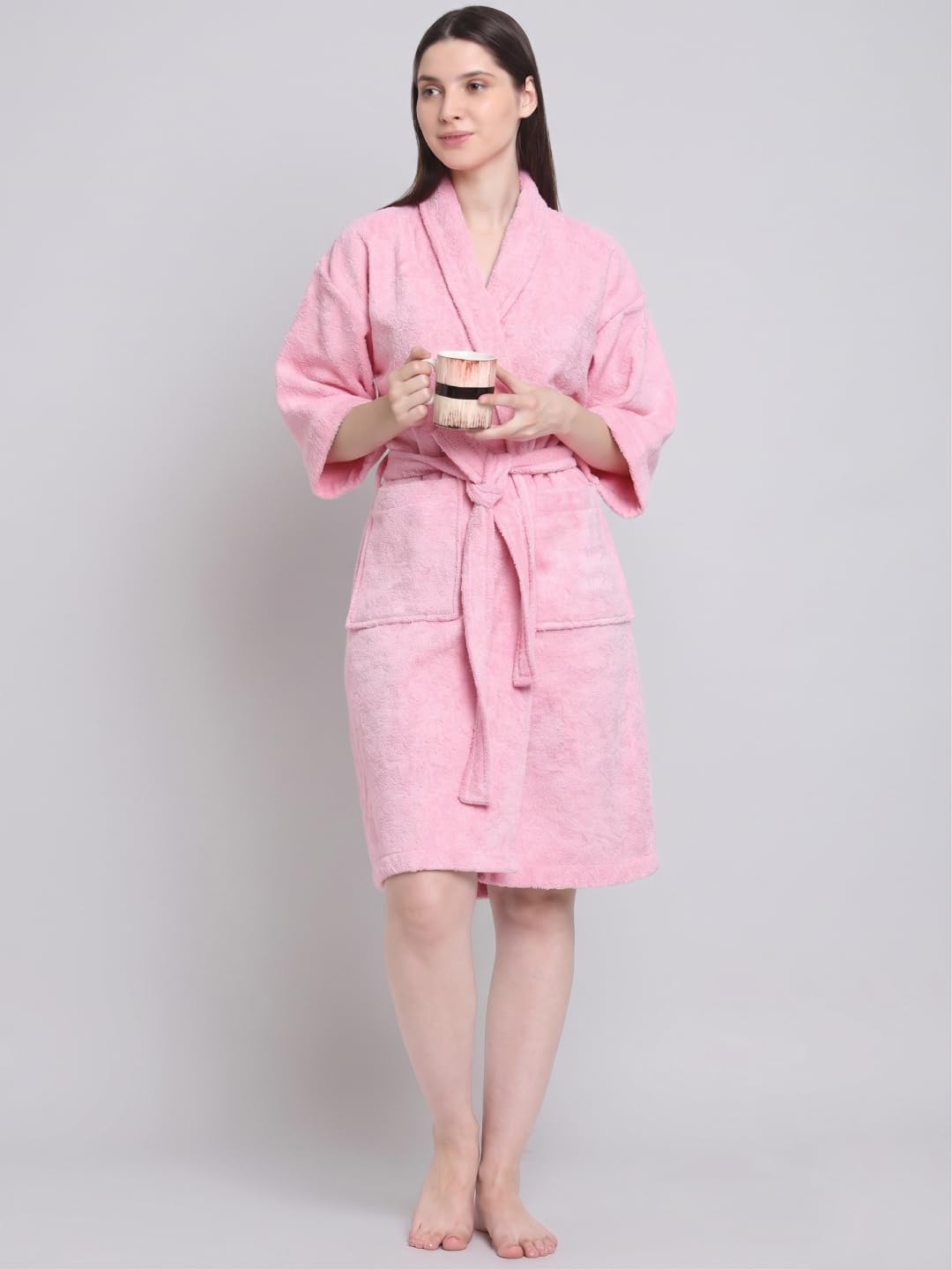 A woman modeling a pink terry cloth bathrobe with a shawl collar, three-quarter sleeves, a self-tie belt, and front patch pockets, set against a plain white background.