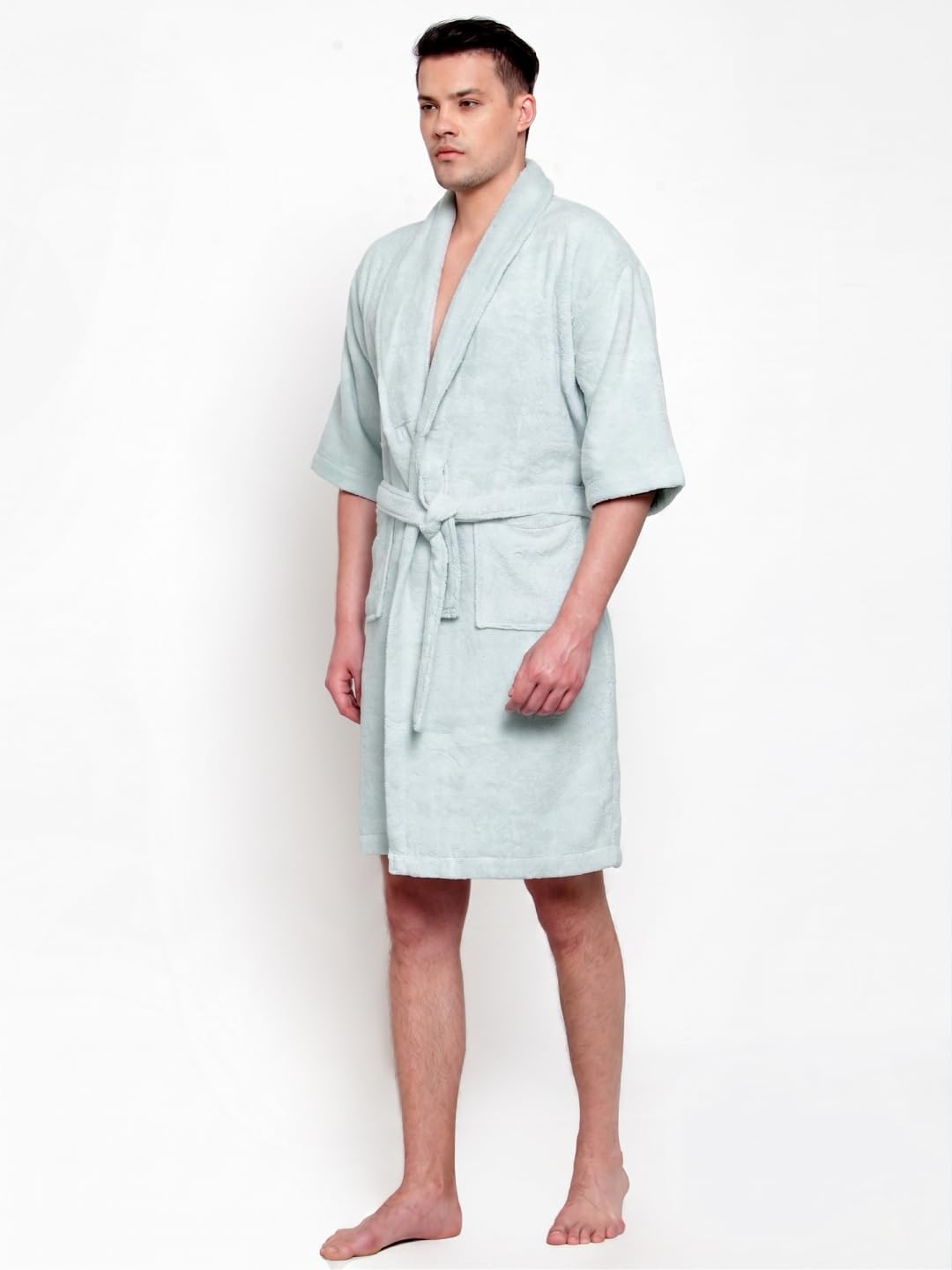 A man modeling a light sage green terry cloth bathrobe with a tie belt and front pockets, standing against a plain white background.