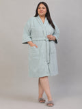 A woman modeling a light sage green bathrobe with a tie belt and front pockets, standing against a neutral gray studio background.
