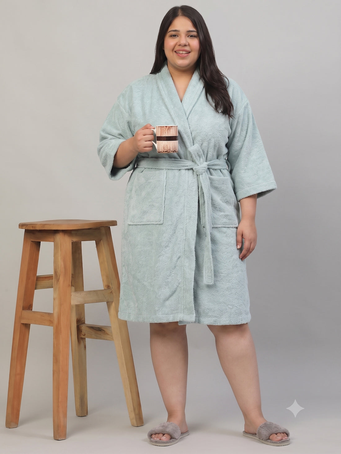 A woman wearing a light sage green terry cloth bathrobe stands next to a wooden stool, holding a small mug. The robe has a tie waist and front pockets, and she is wearing gray slippers against a plain light gray studio background.