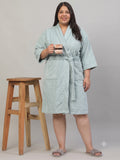 A woman wearing a light sage green terry cloth bathrobe stands next to a wooden stool, holding a small mug. The robe has a tie waist and front pockets, and she is wearing gray slippers against a plain light gray studio background.