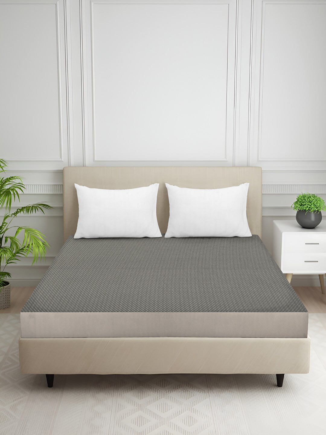 A gray textured bed sheet set with two white pillows on a beige upholstered bed frame, styled in a bright, modern bedroom with a white paneled wall, a side table, and a potted plant.