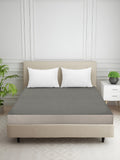 A gray textured bed sheet set with two white pillows on a beige upholstered bed frame, styled in a bright, modern bedroom with a white paneled wall, a side table, and a potted plant.