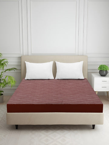 Bed with a red and brown fitted sheet in a room with a white wall and plants.